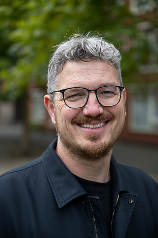 Portretfoto van Wethouder Arjen Kapteijns, en face vanaf de schouders met grote glimlach. Bril met een dun zwart montuur. Foto: Sander Foederer