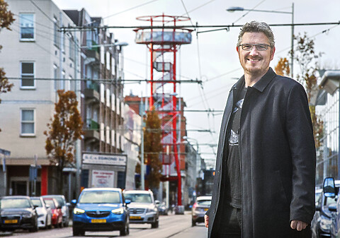 Portretfoto Arjan Kapteijns vanaf heuphoogte, geheel in het zwart gekleed met lange overjas en een grote glimlach. Buiten genomen, met verkeer en de rode panoramatoren op de achtergrond.