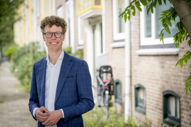 Portretfoto Martijn Balster, gekleed in blauw krijtstreep colbert en wit overhemd zonder stropdas. Een bril met zwart montuur en de handen losjes bij elkaar voor het middel. Foto: Valerie Kuypers