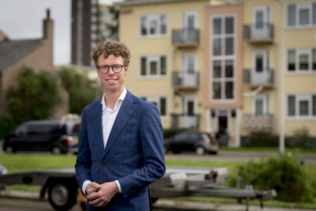 Portretfoto Martijn Balster, gekleed in blauw krijtstreep colbert. Een bril met zwart montuur en de handen in elkaar. Een flatgebouw in Morgenstond op de achtergrond.
