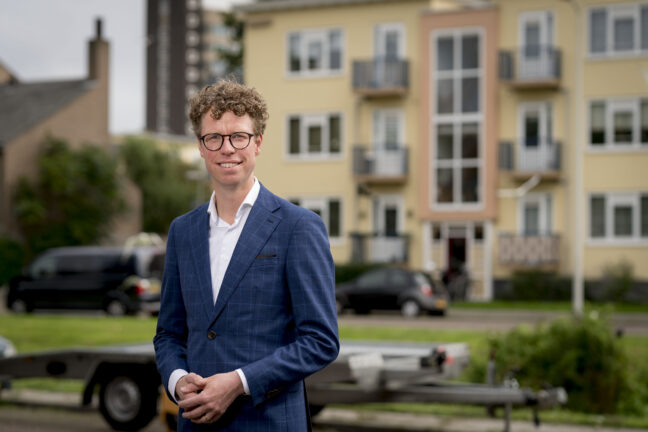 Portretfoto Martijn Balster, gekleed in blauw krijtstreep colbert. Een bril met zwart montuur en de handen in elkaar. Een flatgebouw in Morgenstond op de achtergrond. Foto: Valerie Kuypers