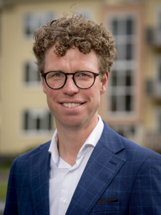 Portretfoto Martijn Balster, close up, gekleed in blauw krijtstreep colbert en wit overhemd zonder stropdas. Een bril met zwart montuur en kalme glimlach. Foto: Valerie Kuypers