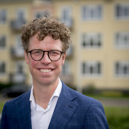 Portretfoto Martijn Balster, close up, gekleed in blauw krijtstreep colbert en wit overhemd zonder stropdas. Een bril met zwart montuur en een grote glimlach. Foto: Valerie Kuypers