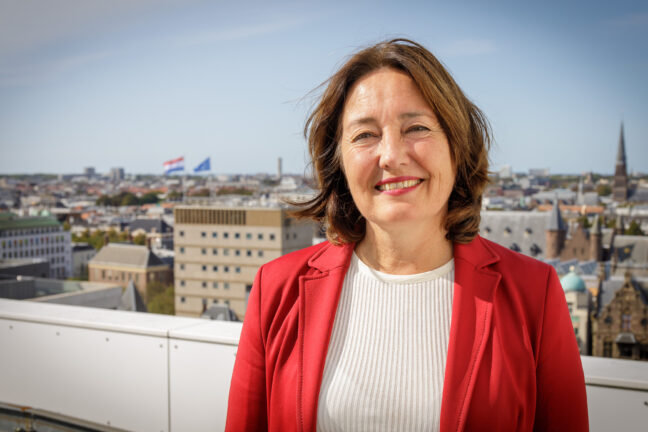 Portretfoto van Saskia Bruines, en face, genomen vanaf het dak van het stadhuis. Saskia draagt een bessenrood jasje op een wit shirt en glimlacht met de kin iets omhoog.