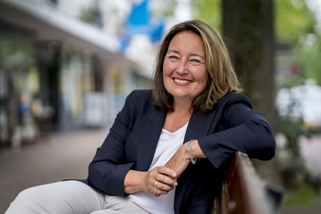 Portretfoto van Saskia Bruines, zittend op een bankje, de schouders gedraaid naar de camera, een elleboog op de rugleuning. Saskia draagt een donkerblauw jasje op wit shirt, een spontane glimlach en toegeknepen ogen. (foto: Valerie Kuypers)