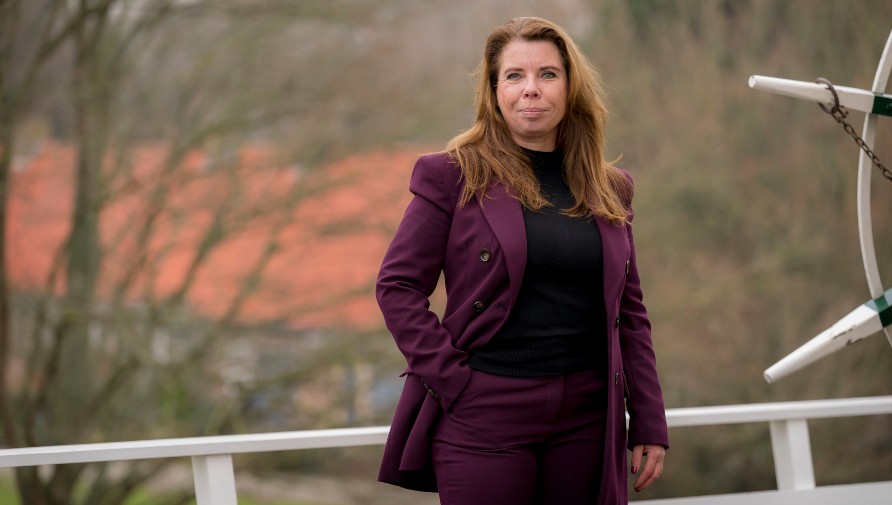 Portretfoto van Corrine den Heijer in paars broekpak en zwarte coltrui. Ze heeft 1 hand in de zak.