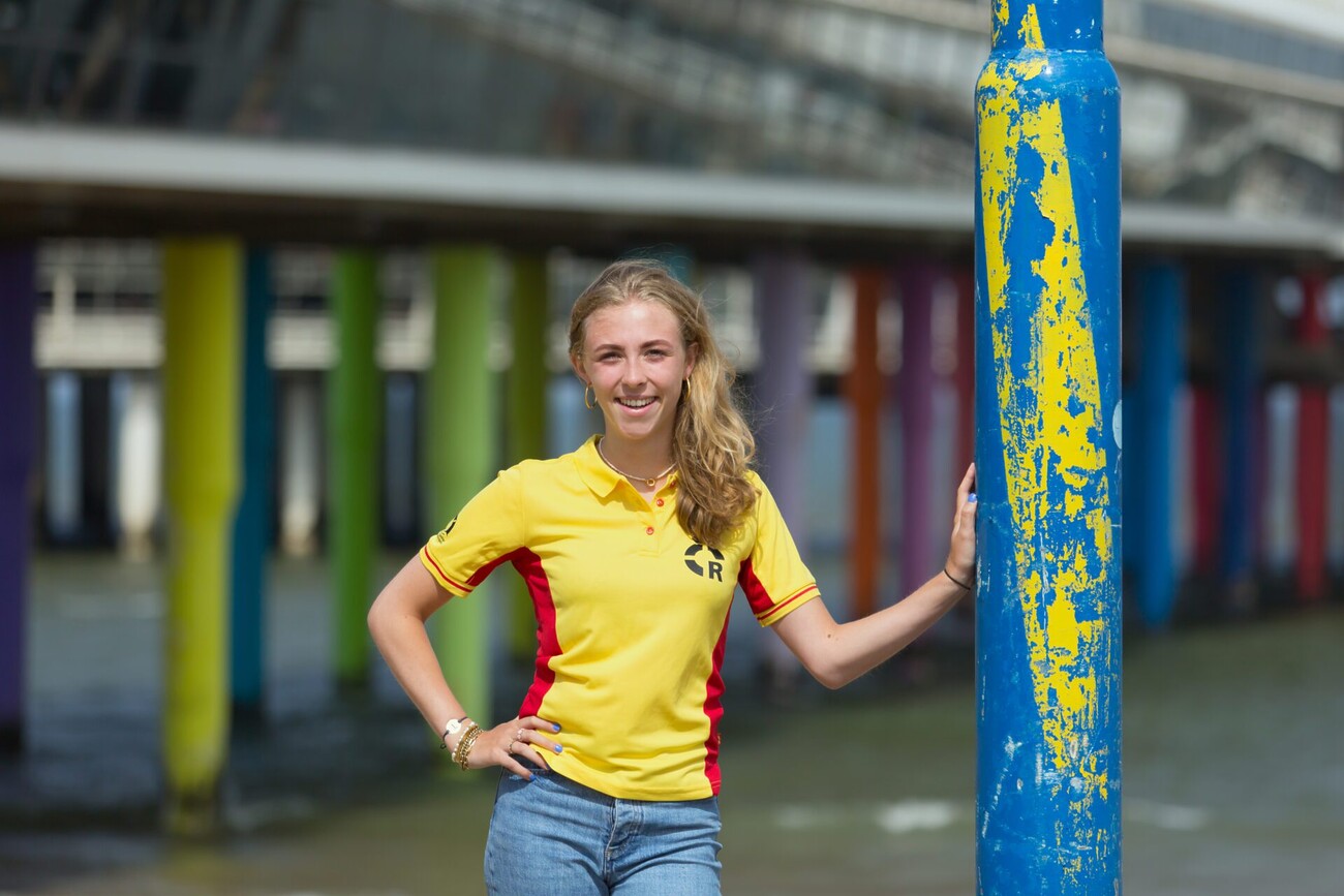 Lifeguard Titia staat met een hand op de heup in fel geel t-shirt met 2 rode strepen aan de zijden tegen een blauw gekleurde paal geleund. De pijlers van de pier liggen achter haar.