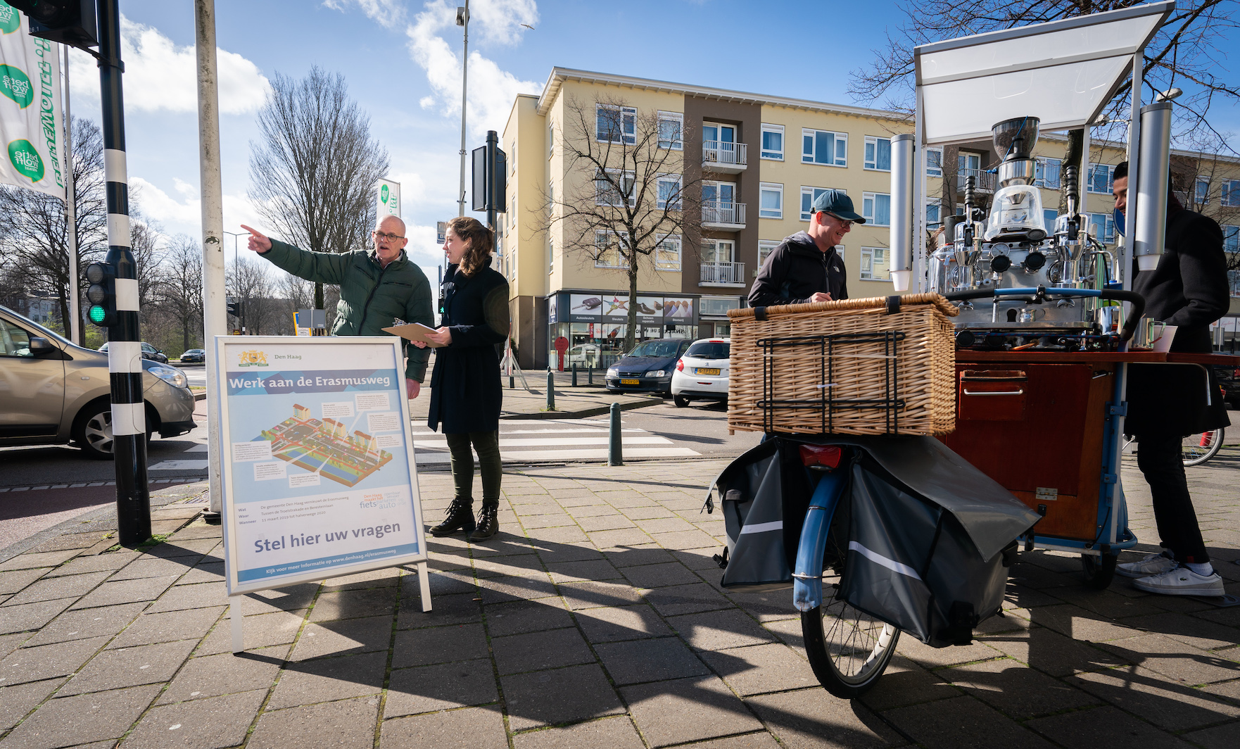 Een hoek op een kruispunt met de ERasmusweg. Een bord kondigt werkzaamheden aan. Een man, kaal wijst met zijn hand naar rechts, een jonge vrouw met clipboard staat naast hem. Rechts staat een verrijdbare koffiekraam.