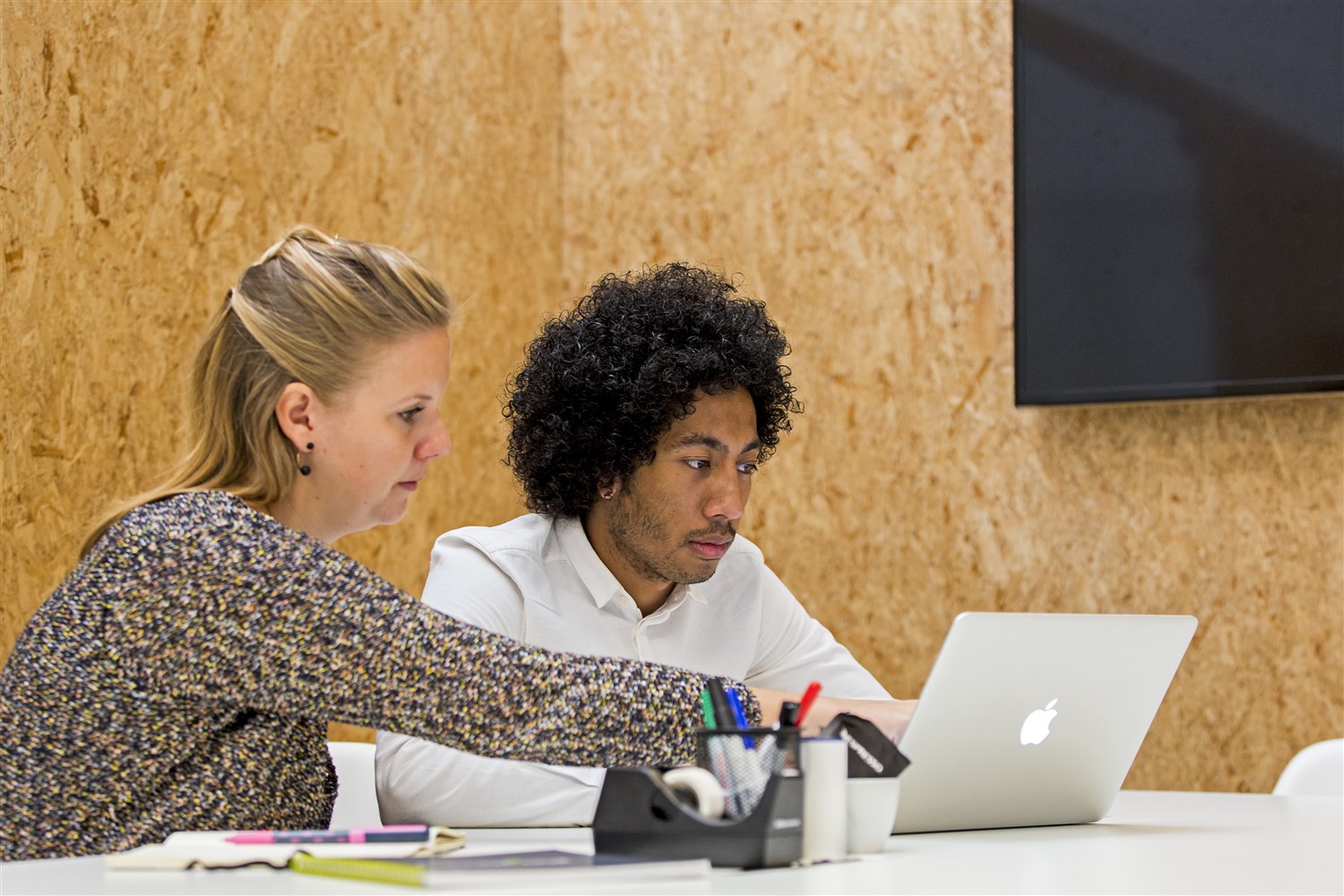 Een vrouw, blond, wijst iets aan op de laptop van een jongen, kort krullend zart haar. Beide zijn in de twintig.