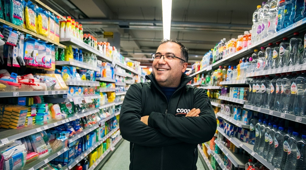 Portret van Yakup Uysal in de gangen van zijn Coopsupermarkt, de armen over elkaar en grote glimlach. De huishoudelijke producten links, de flessen (fris)drank rechts.