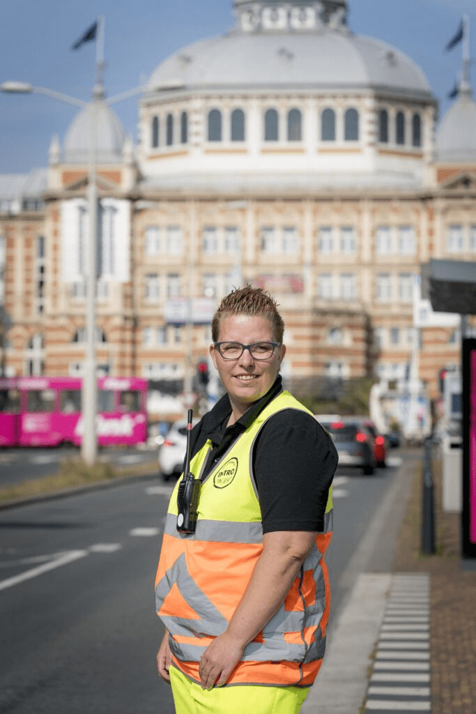 Verkeersregelaar naomi staat in reflecterend tenue bij een bushalte met een glimlach en toegeknepen ogen door tegenlicht. Het Kurhaus ligt vlak achter haar.