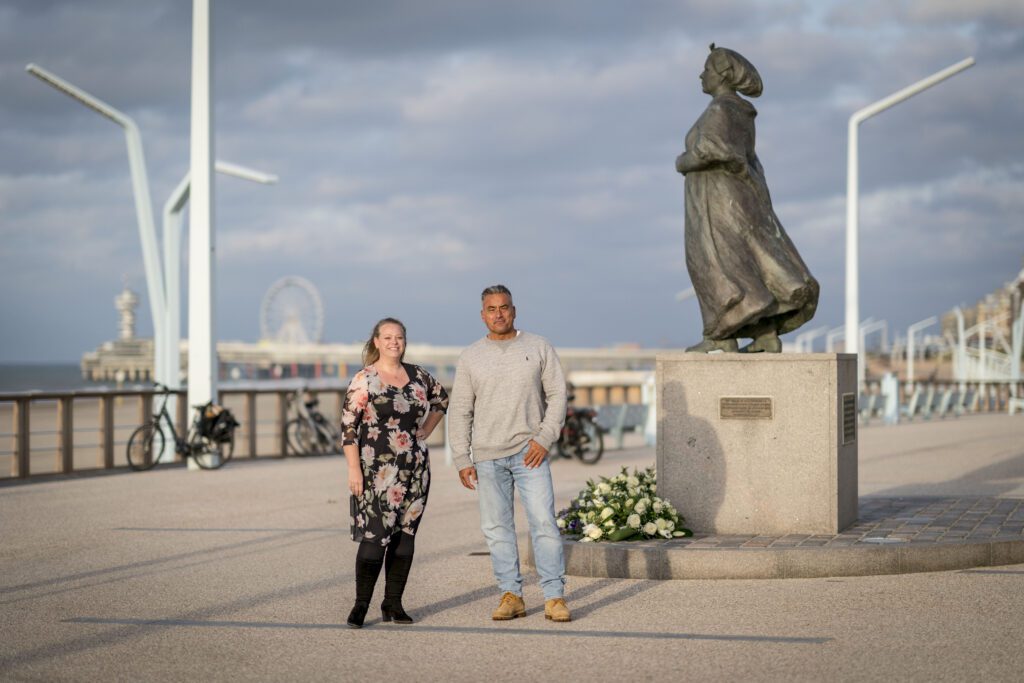 Roos in gebloemde jurk op zwarte achtergrond en Lars in lichtgrijze trui en lichtblauwe jeans. Beide staan in avondzon bij het standbeeld van de Vissersvrouw van Scheveningen.