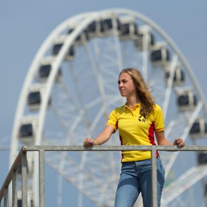 Portret, klein, van Titia in fel geel lifeguard shirt met het reuzenrad van de pier op de achtergrond.