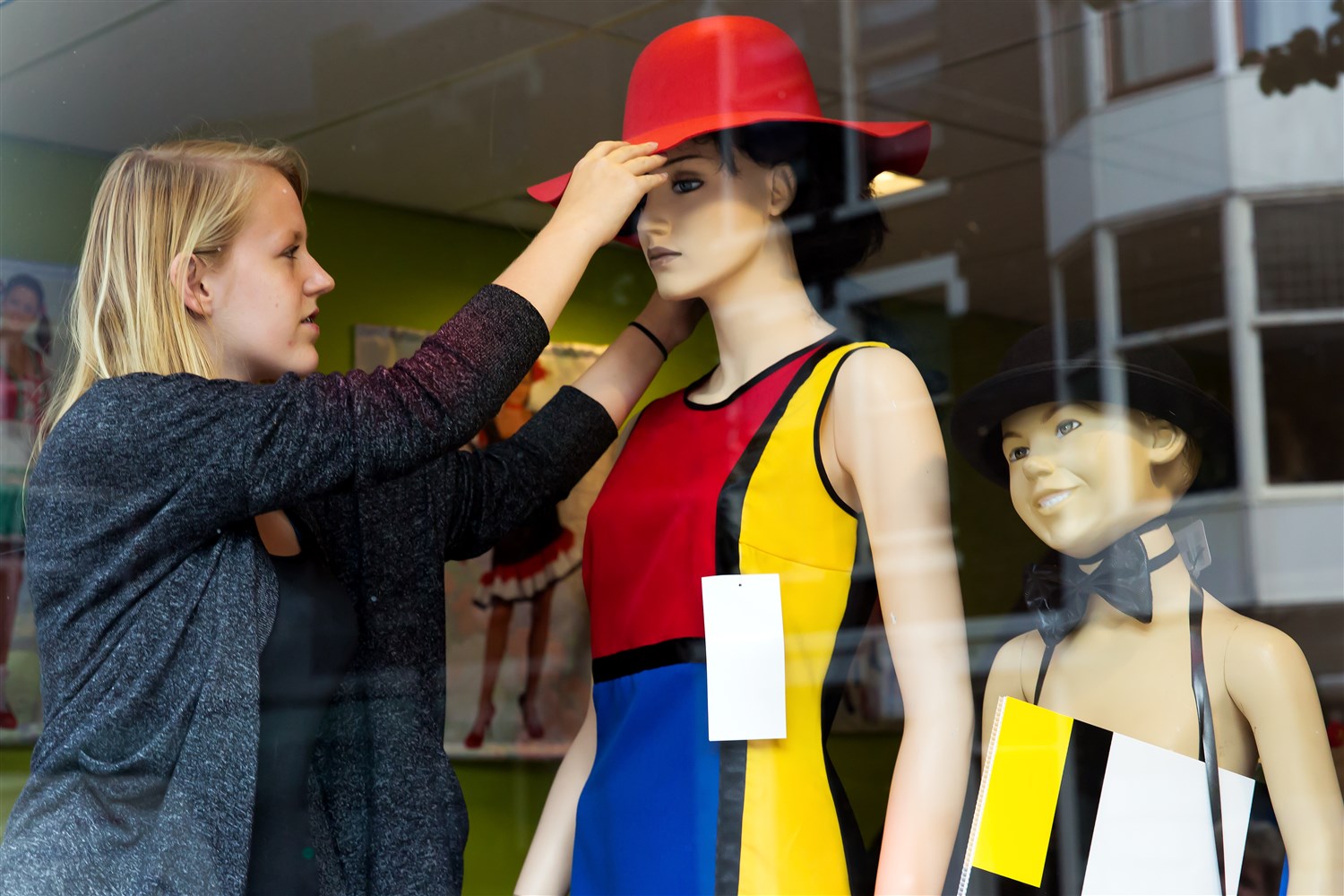 Een jonge, blonde vrouwe zet een rode hoed op een etalagepop die gekleed is in een mondriaan, mouwloos jurkje.