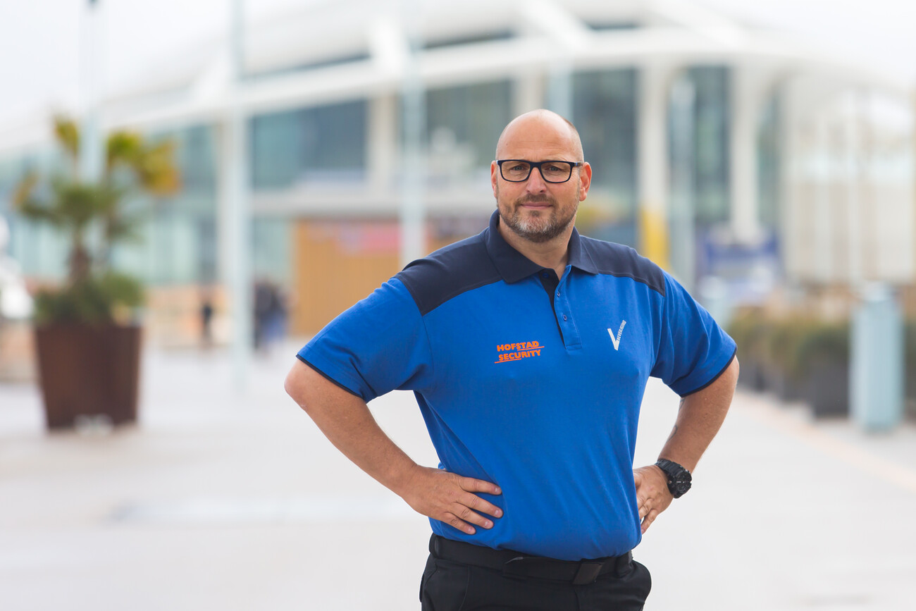 Cees poseert in midblauwe poloshirt met logo van Hofstad Security op de borst en de handen in de zijden.