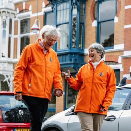 een oudere man en vrouw lopen lachend in fel oranje jasjes over straat