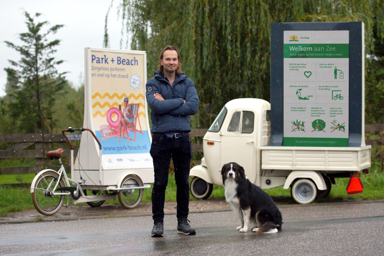 Dimitri, in blauw donsjack, poseert met hond voor een abribike ('Park + Beach') en abricar ('Welkom aan zee').