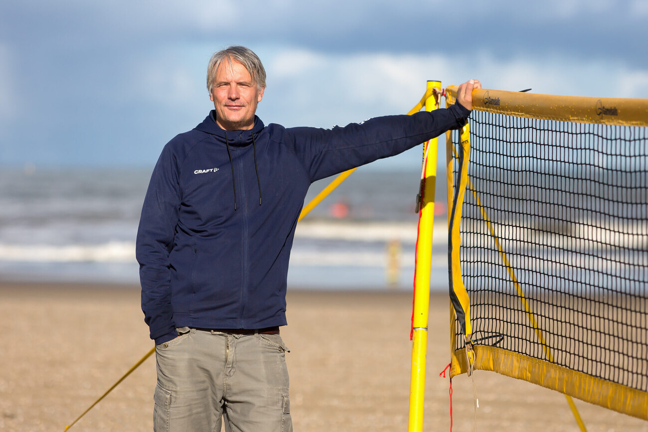 Rick in donkerblauw trainingsjack leunt op het strand tegen een fel geel volleybalnet