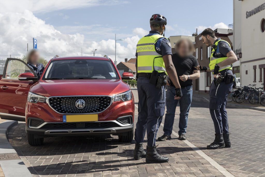 2 parkeerhandhavers houden een rode auto aan. De bestuurder en bijrijder zijn onherkenbaar gemaakt.