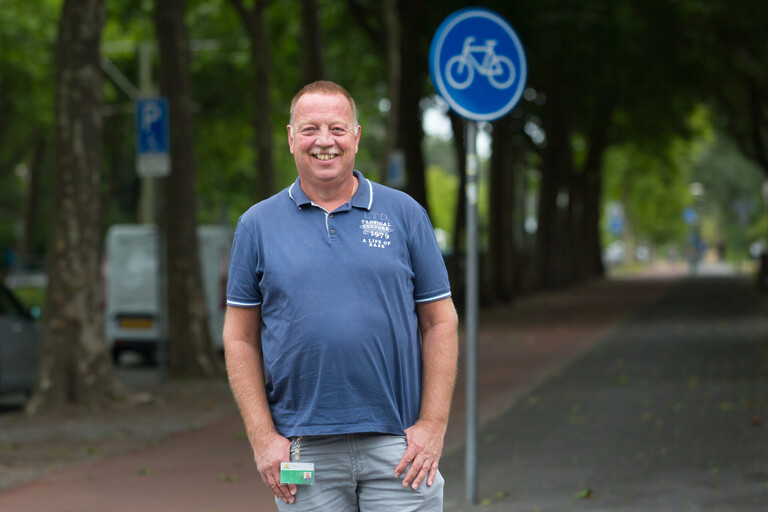 Peter poseert buiten met duimen in de zakken in blauwe polo en lichtgrijze broek. Zijn gemeentepas hangt aan zijn broek. Hij heeft een brede glimlach.