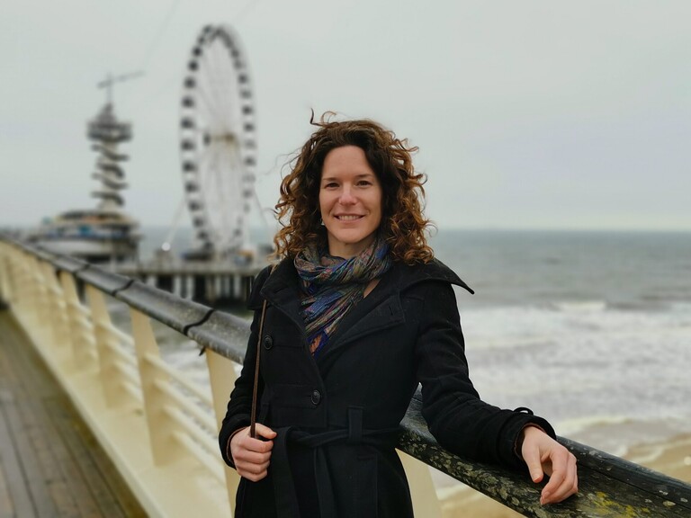 Saskia leunt in zwarte wollen jas en sjaal tegen de balustrade van de pier.