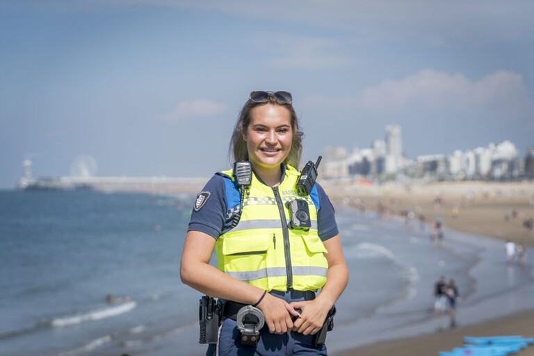 Tara poseert in uniform en beveiligingsvest met het strand en de zee op de achtergrond.