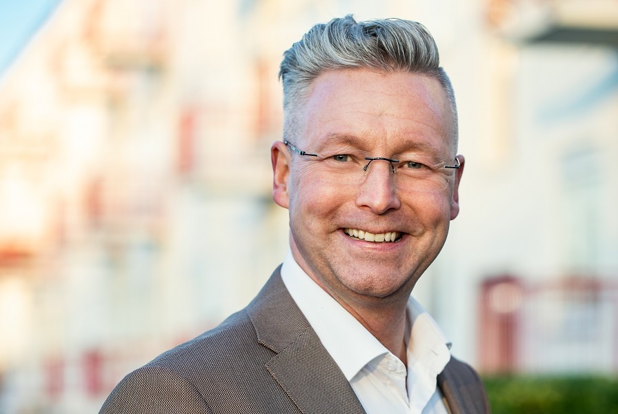 Portret van Rick Theunissesn, vanaf de schouders. Lichtgrijs pak met hoog wit boord, een montuurloze bril en kort grijs haar.