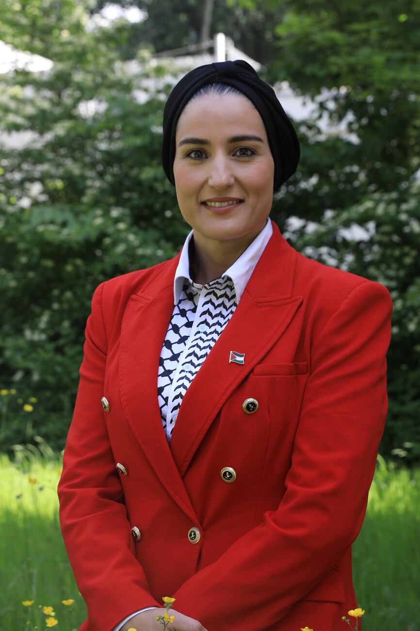 Jonge vrouw met strak om het hoofd gebonden zwarte hoofddoek en gekleed in een felrode blazer glimlacht naar de camera in een parkachtige omgeving