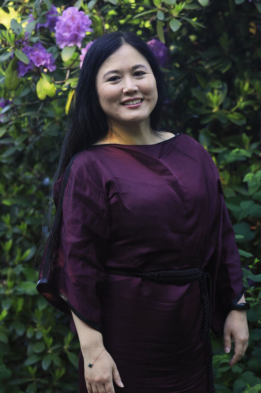 Glimlachende vrouw met zwart lang haar, gekleed in een auberginekleurige jurk, poseert voor een rodondendron in bloei