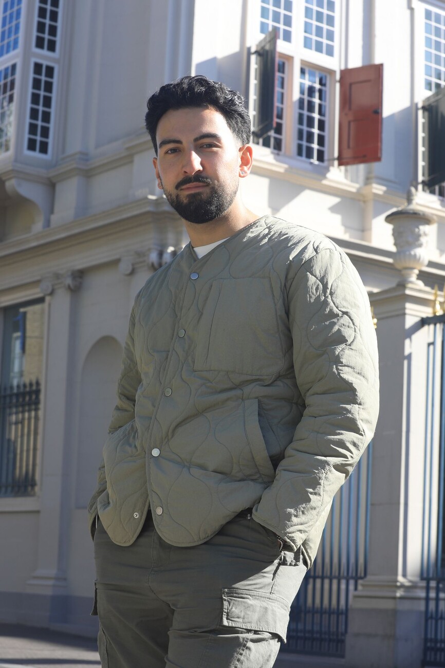Man met baard, gekleed in een olijgroen gewatteerd jasje, poseert met zijn handen in zijn broekzakken voor een herenhuis