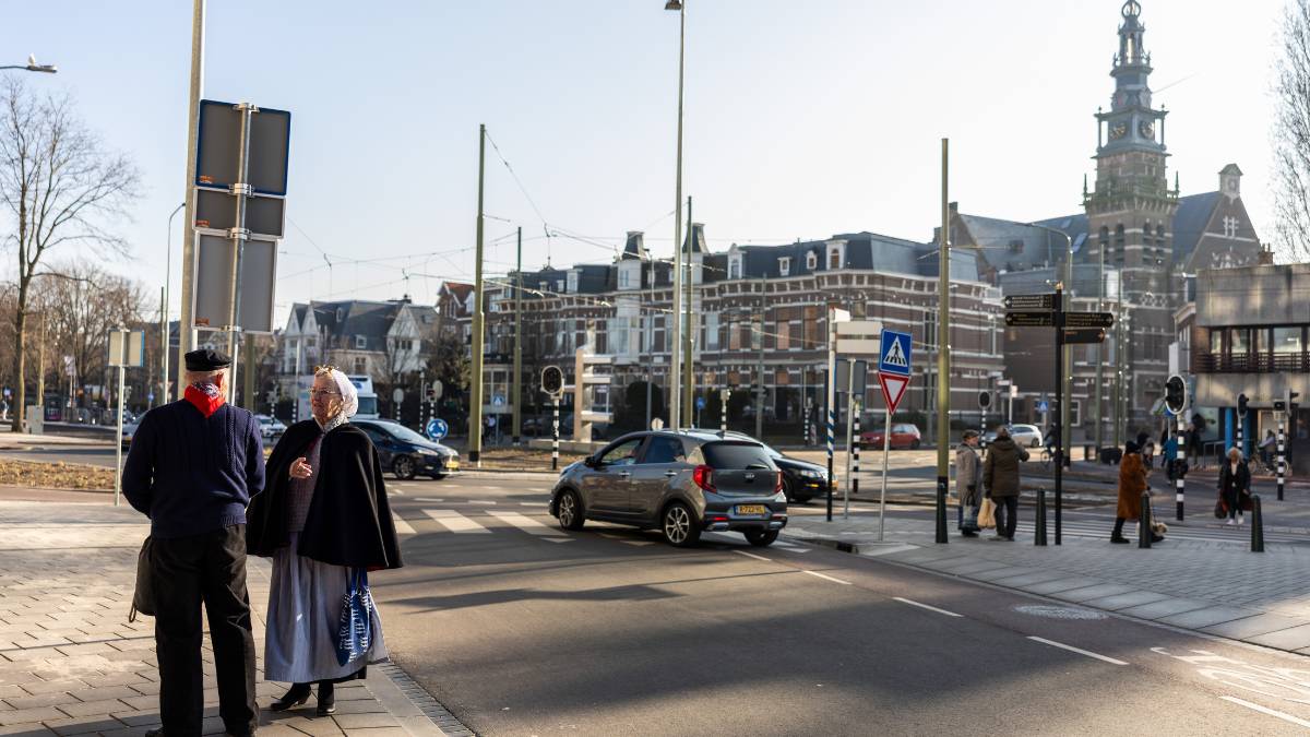 Twee mensen in traditionele klederdracht steken een straat over in Scheveningen-Dorp, met op de achtergrond historische panden.