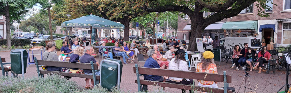 Het Valkenbosplein. Bewoners zitten op bankjes en stoelen en praten met elkaar.