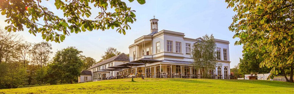 Villa Ockenburgh vanaf een afstand op de foto. Omgeven door groen gras en een terras met parasollen.