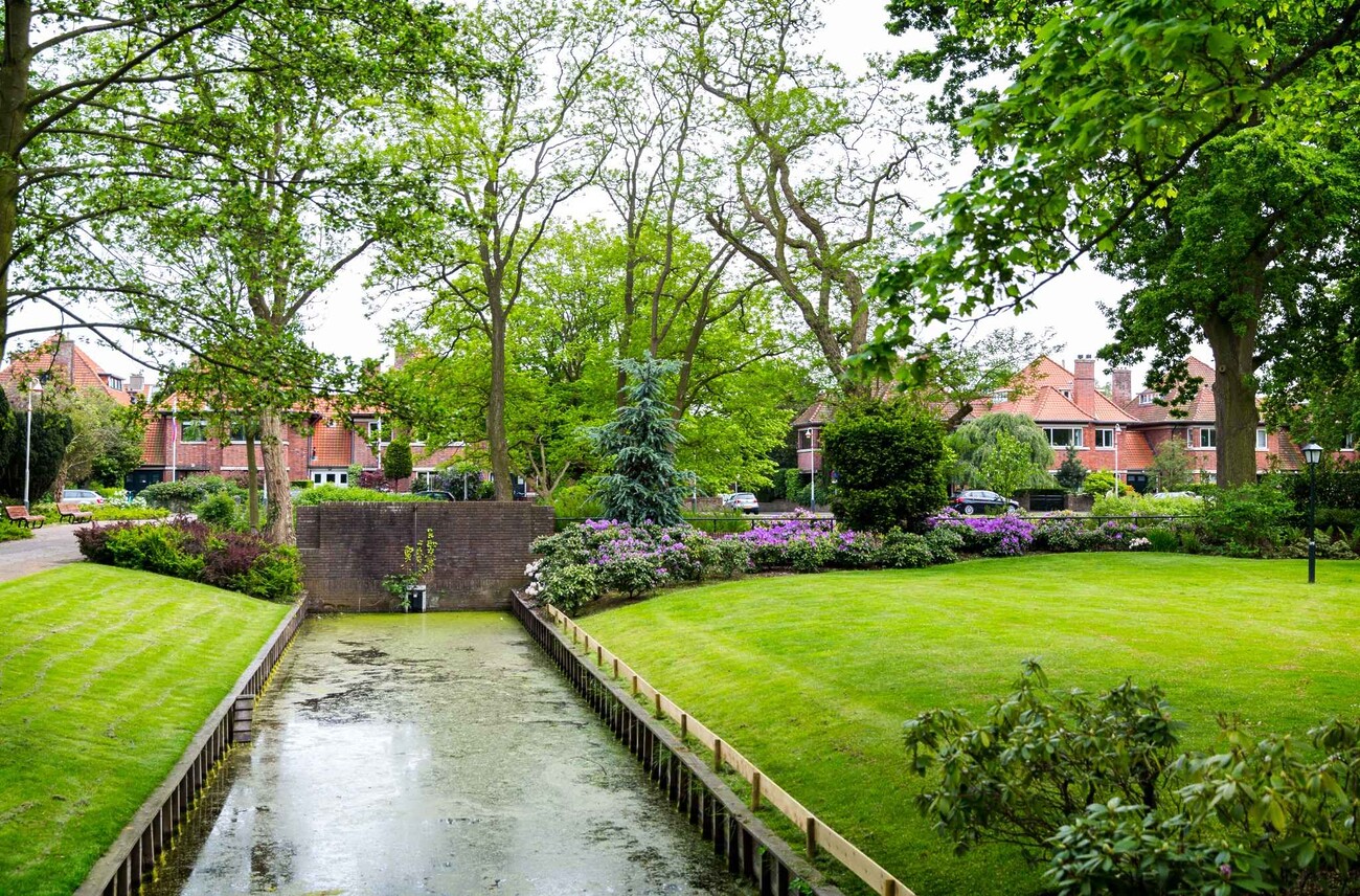 Een groen park met gras en bloemen. In het midden ligt een smal kanaal. Aan de rand staan bomen en struiken. Op de achtergrond zie je huizen met rode daken. Het is een rustige wijk.