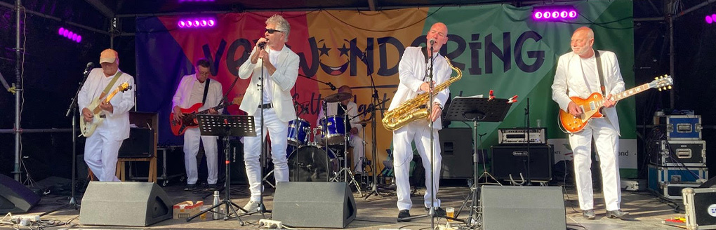 Bandleden maken muziek op een podium. 2 op (bas)gitaar, 1 speelt saxofoon en 1 zingt.