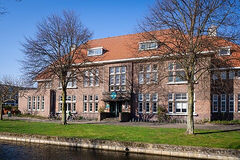 Bakstenen schoolgebouw met rood dak. Veel ramen aan de voorzijde. Voor het gebouw ligt een gracht. Grasveld met bomen en fietsen.