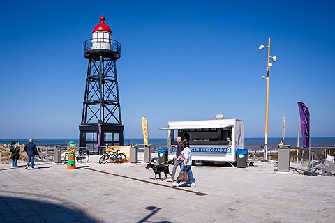 Lichttoren aan de boulevard. Snackkar met mensen ervoor. Felle zon en blauwe lucht. Zee in de verte. Borden, vlaggen en fietsen aanwezig.
