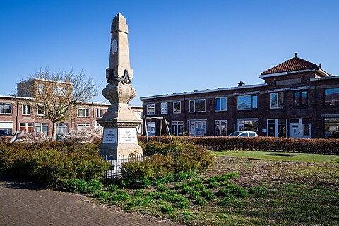 Oud gedenkteken midden op een plein. Omringd door lage struiken. Rijtjeshuizen op de achtergrond. Blauwe lucht en zonlicht. Kleine speelplek ernaast.