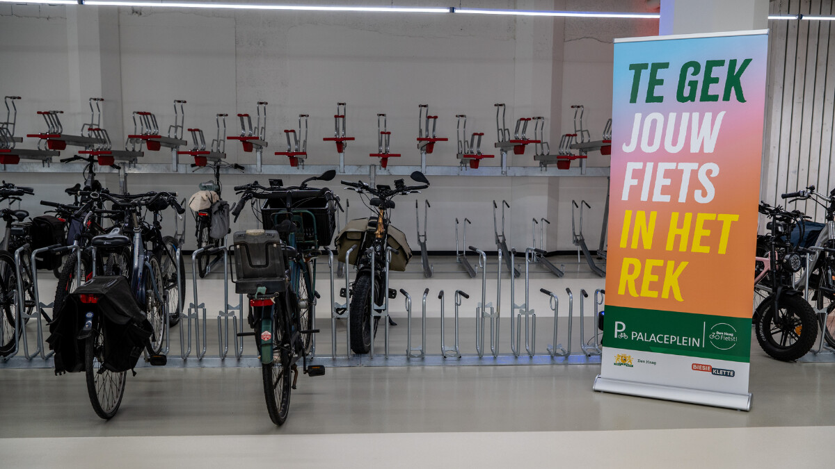 Fietsen staan geparkeerd in fietsenstalling 'Palaceplein'. Er staat een kleurrijke banner met 'Te gek, jouw fiets in het rek'.