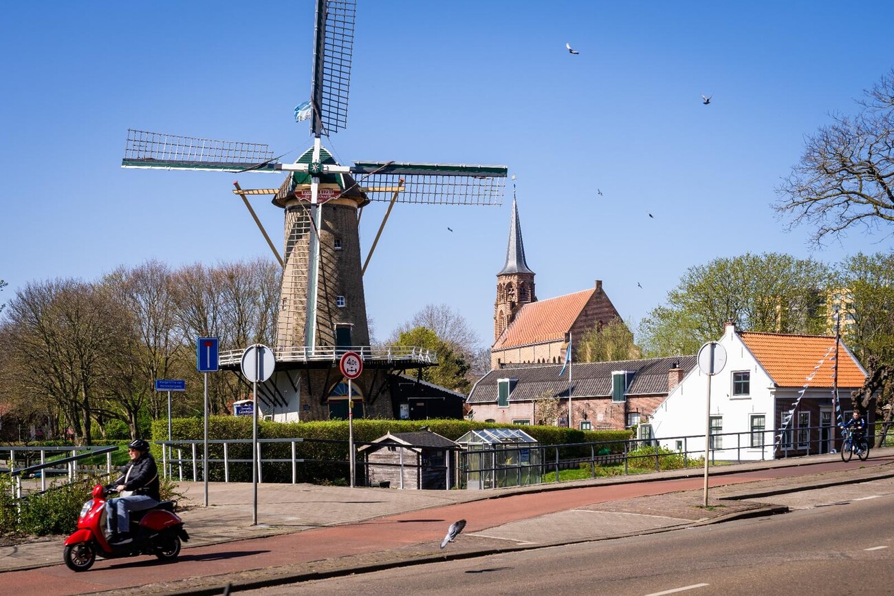 Windmolen naast een oude kerk. Rode scooter rijdt voorbij. Blauwe lucht en vogels in de lucht.