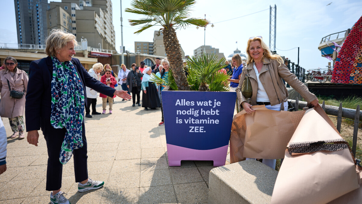 Onthulling van het nieuwe Pierplein. De stadsdeeldirecteur wijst naar een tekst op een plantenbak: 'Alles wat je nodig hebt is vitamine ZEE'. Het is een zonnige dag.