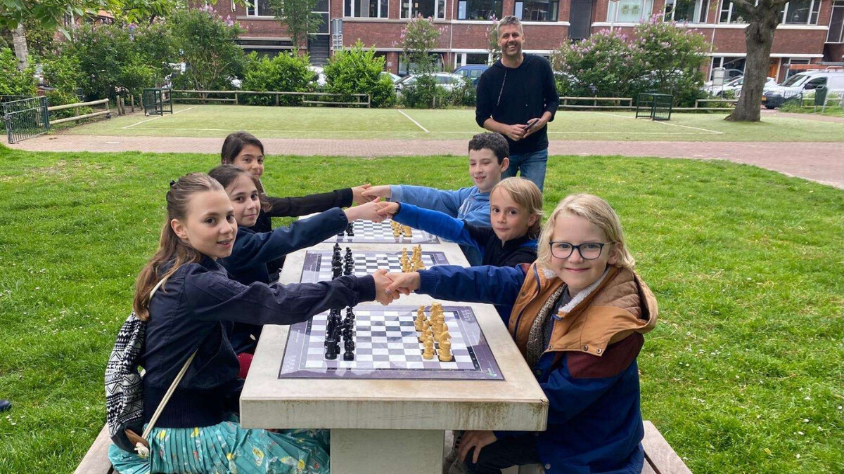 Kinderen zitten bij schaaktafels op een grasveld en schudden elkaar de hand.