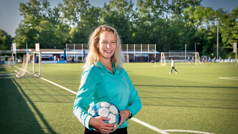 Marjolein Kool op een voetbalveld in Loosduinen. Ze heeft een bal in haar hand en kijkt lachend naar de camera.