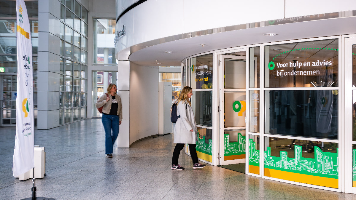 Twee personen lopen in een hal naar de ingang van het Ondernemersportaal. Op de glazen deur staat “Voor hulp en advies bij ondernemen”