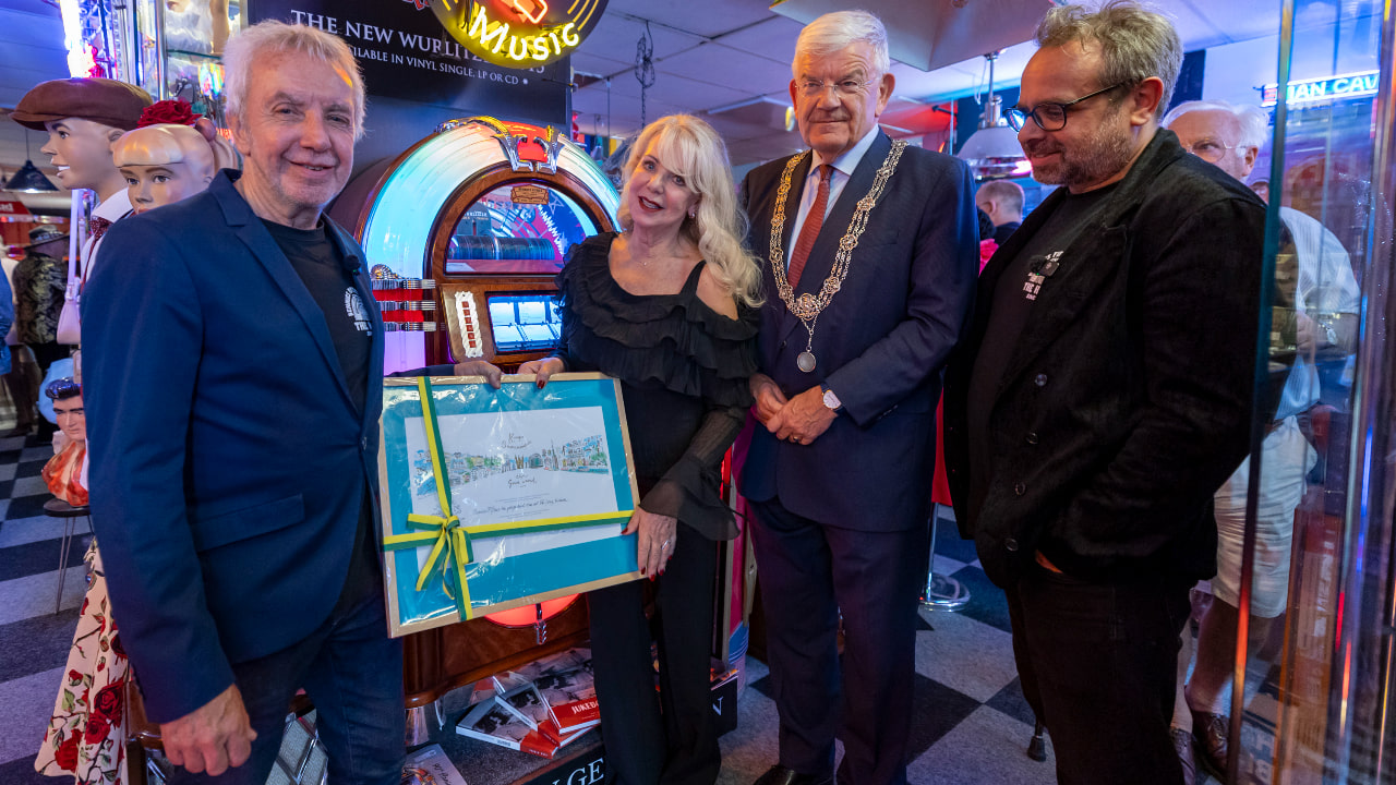 4 personen staan in een winkel met retro en muziekartikelen rond een jukebox. 1 persoon houdt een ingelijst document met een groen‑gele strik vast, terwijl de anderen ernaast staan. Op de achtergrond zijn neonlichten, etalagepoppen en muziekgerelateerde decoratie te zien.
